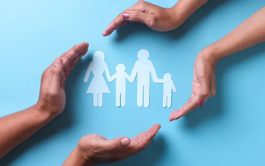 A paper cut-out family protected by four hands on a blue background