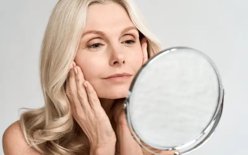 Middle-aged woman examining skin in mirror for signs of aging and skincare results